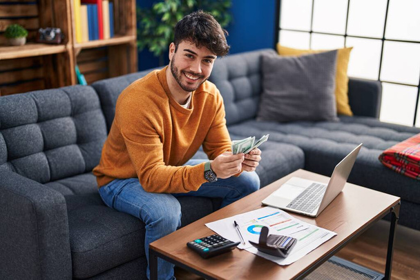 Young hispanic man using laptop holding dollars sitting on sofa at home - Фото, зображення