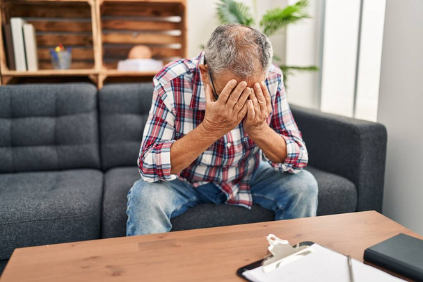 Homme âgé aux cheveux gris patient stressé avec les mains sur le visage assis sur le canapé à la clinique de psychologie - Photo, image