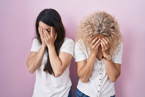 Mother and daughter standing together over pink background with sad expression covering face with hands while crying. depression concept.  - Photo, image
