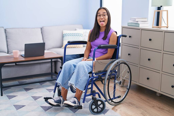Νεαρή Ισπανίδα που κάθεται σε αναπηρικό καροτσάκι στο σπίτι και βγάζει τη γλώσσα της χαρούμενη με την αστεία έκφραση. έννοια συναισθήματος.  - Φωτογραφία, εικόνα