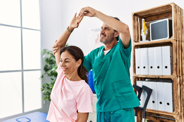 Мужчина и женщина среднего возраста улыбаются уверенно растягивая руку, проводя сеанс реабилитации в клинике физиотерапии - Фото, изображение