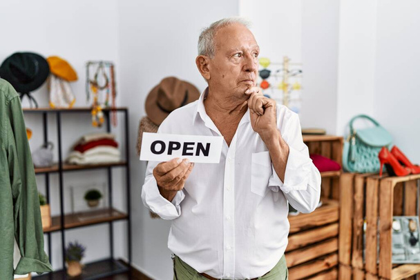 Senior man holding banner with open text at retail shop serious face thinking about question with hand on chin, thoughtful about confusing idea  - Foto, imagen