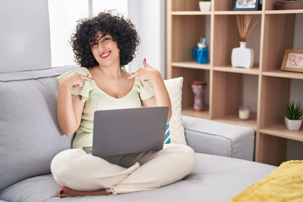 Junge brünette Frau mit lockigem Haar mit Laptop, die zu Hause auf dem Sofa sitzt und selbstbewusst lächelt und stolz und glücklich mit den Fingern zeigt.  - Foto, Bild