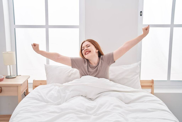 Fiatal vörös hajú nő felébred kinyújtott karokkal a hálószobában - Fotó, kép