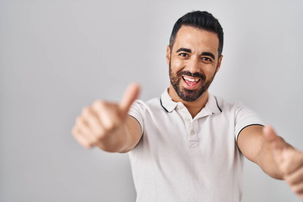 Young hispanic man with beard wearing casual clothes over white background approving doing positive gesture with hand, thumbs up smiling and happy for success. winner gesture.  - Foto, Imagem