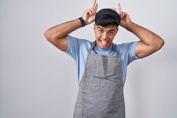 Hispanic young man wearing apron over white background posing funny and crazy with fingers on head as bunny ears, smiling cheerful  - Photo, Image