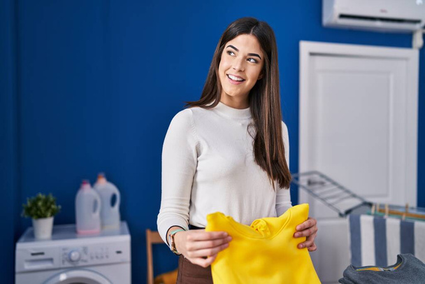 Nuori kaunis latino nainen hymyilee luottavainen taitto vaatteita pesutupa - Valokuva, kuva