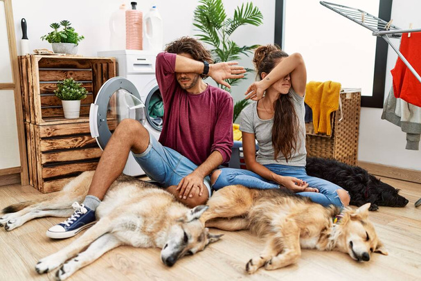 Young hispanic couple doing laundry with dogs covering eyes with arm, looking serious and sad. sightless, hiding and rejection concept  - Photo, Image