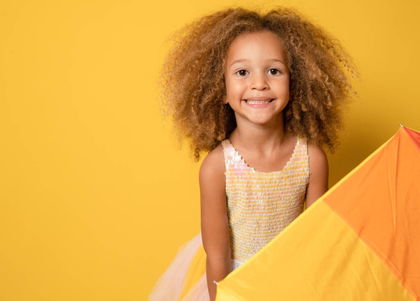 Close up portret Klein kind meisje met krullend haar staan onder kleurrijke paraplu geïsoleerd op gele achtergrond. - Foto, afbeelding