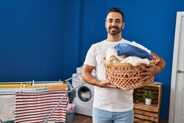 Junger hispanischer Mann mit Bart und Wäschekorb, der positiv und glücklich im Stehen aussieht und mit einem selbstbewussten Lächeln, das Zähne zeigt, lächelt  - Foto, Bild