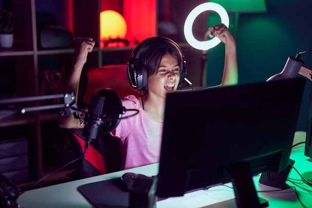 Adorable hispanic girl streamer playing video game with winner expression at gaming room - Foto, Imagem
