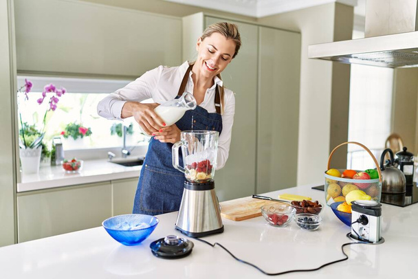 Joven mujer rubia sonriendo confiada vertiendo leche en la licuadora en la cocina - Foto, imagen