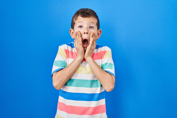 Young caucasian kid standing over blue background afraid and shocked, surprise and amazed expression with hands on face  - Photo, Image