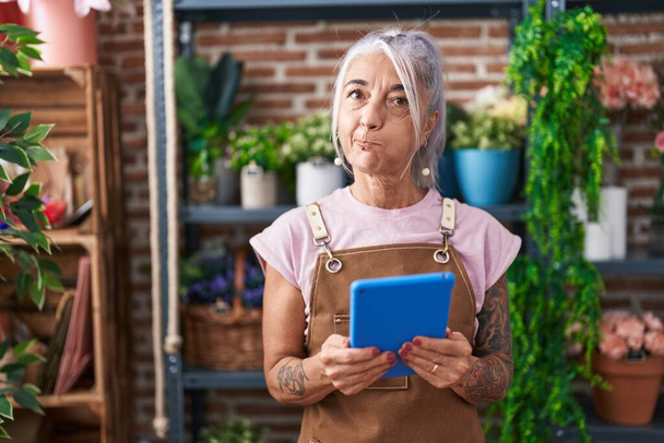 Femme d'âge moyen avec des tatouages travaillant à la boutique de fleuriste avec tablette gonflant les joues avec un visage drôle. bouche gonflée d'air, attrapant l'air.  - Photo, image