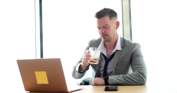 Empresario está sosteniendo un vaso de whisky y mirando a la computadora portátil debido al estrés del trabajo duro debido a las malas condiciones económicas. Alcoholismo en el lugar de trabajo - Imágenes, Vídeo