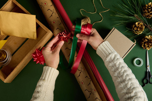 View from above of womans hands, hold red and green satin decorative tapes, a tied shiny bow over wrapping gift paper, packing presents on a surface with ornaments. Diy. Creative art idea. Handwork - Foto, imagen