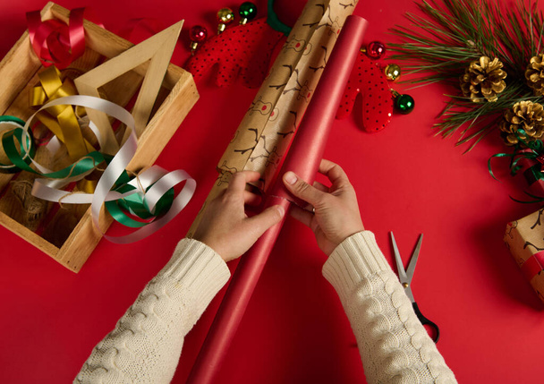 Draufsicht weiblicher Hände, die Geschenkpapier und dekorative Schleifen aufheben, während sie Weihnachts- oder Neujahrsgeschenke auf roter Oberfläche verpacken. Weihnachtsvorbereitungen. Tag des Boxens. Frohe Winterferien. Feier - Foto, Bild