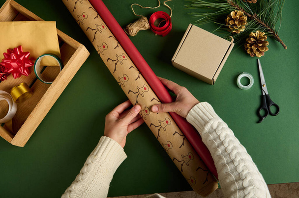 トップビューの手は、染料プレゼントのための装飾材料と緑の背景に包装紙を保持しています。新年とクリスマスの準備。手芸だ。工芸品だ。贈り物を詰めろ。ボクシングの日。魔法の気分 - 写真・画像