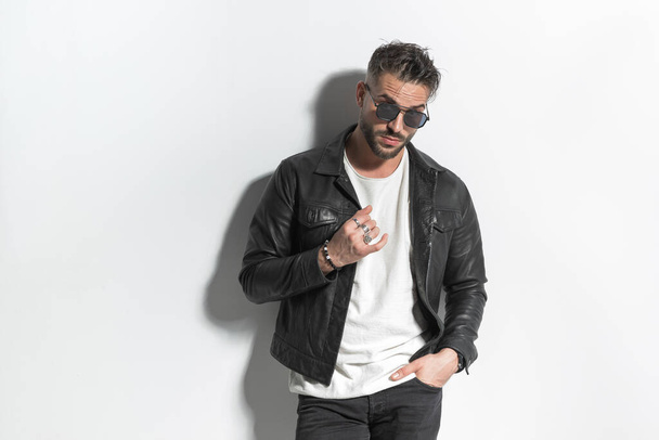 sexy latin man with beard and wet brunette hair posing with hand in pocket while fixing leather jacket and looking away in front of grey wall - Фото, изображение