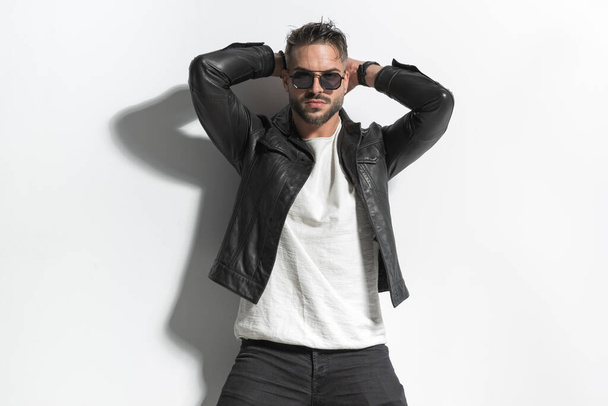 sexy uomo di moda in giacca di pelle con occhiali da sole che si tengono le mani dietro la testa e posa in modo fresco di fronte al muro grigio in studio - Foto, immagini