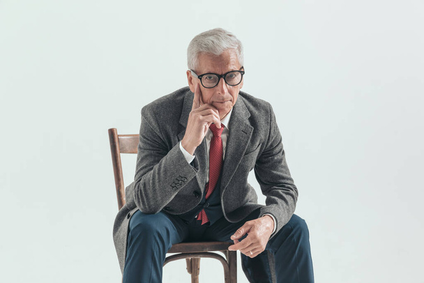 homem pensativo com cabelos grisalhos tocando rosto e pensamento, posando com cotovelos nos joelhos enquanto sentado em fundo cinza no estúdio - Foto, Imagem