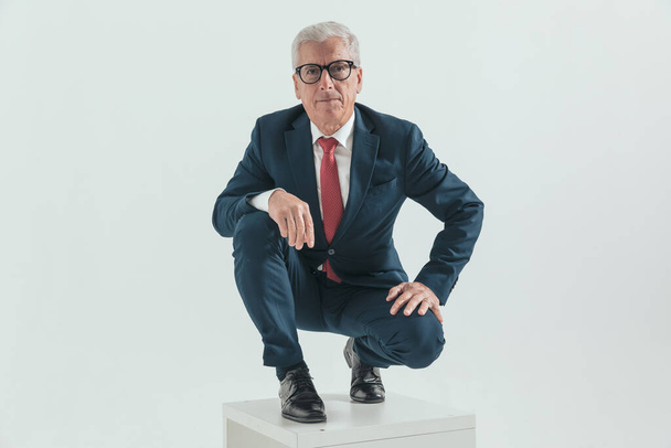 elegante viejo hombre de negocios con gafas sosteniendo los brazos en las rodillas y agachándose delante de fondo gris en el estudio - Foto, Imagen