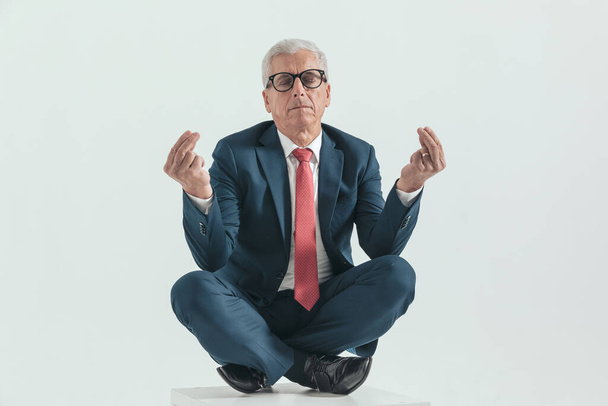 old man in his 60s in a turkish pose holding elbows on knees, closing eyes and meditating in front of grey background - Photo, Image