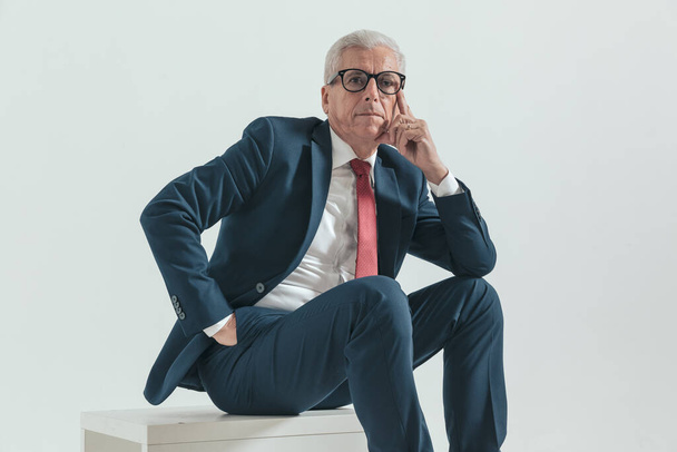 seated elegant man with grey hair touching face and thinking while posing with hand in pocket and elbow on knee in front of grey background in studio - Photo, Image