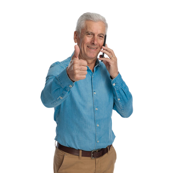 viejo guapo hablando por teléfono, siendo feliz, sonriendo y haciendo pulgares hacia arriba signo delante de fondo blanco en el estudio - Foto, Imagen