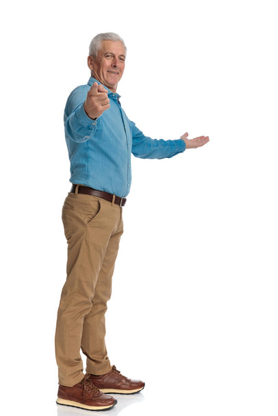 gelukkige oude man wijzende vinger en presenteren aan de kant terwijl glimlachen en staan in de rij voor witte achtergrond in de studio - Foto, afbeelding