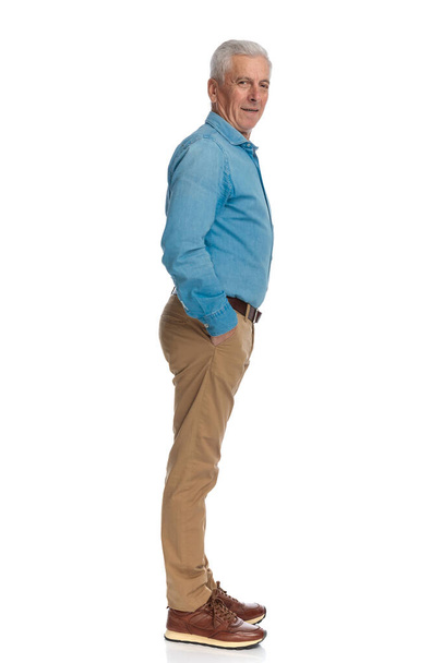 πλαϊνή άποψη του casual γέρος με τα χέρια στις τσέπες στέκεται στην ουρά και περιμένει στο λευκό φόντο στο στούντιο - Φωτογραφία, εικόνα