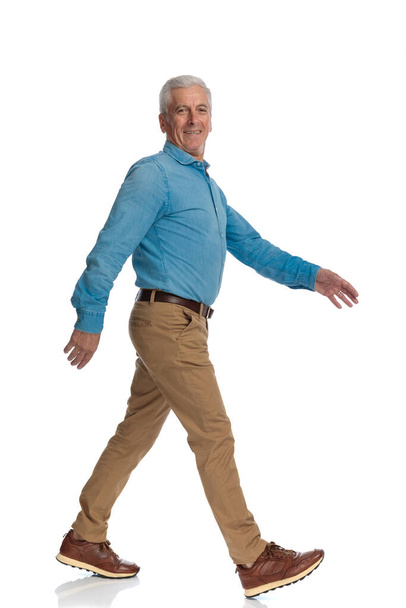 vista lateral de hombre viejo feliz con zapatillas sonriendo y caminando delante de fondo blanco en el estudio  - Foto, imagen