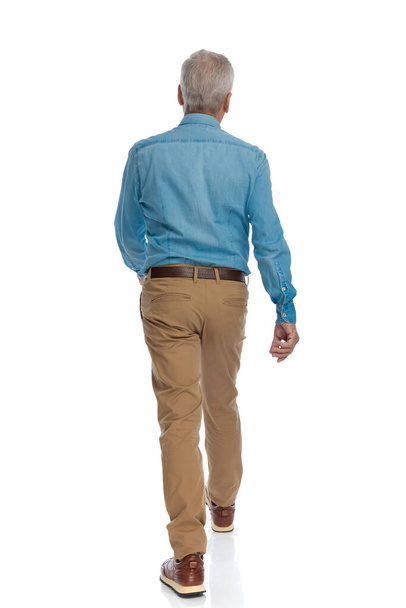 widok z tyłu przypadkowy starzec w dżinsowej koszuli i spodnie chino wchodzenie przed białym tle w studio - Zdjęcie, obraz