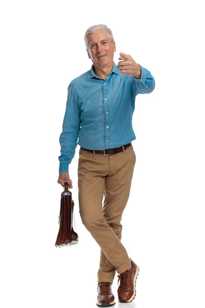 Evrak çantalı mutlu yaşlı adam, stüdyoda beyaz bir arkaplan önünde dikilirken seçiyor ve gülümsüyor. - Fotoğraf, Görsel