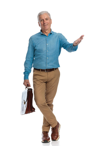 šedesátiletý šedovlasý prošedivělý muž s kufrem zvednutou paží a gestem, zatímco stojí před bílým pozadím ve studiu - Fotografie, Obrázek