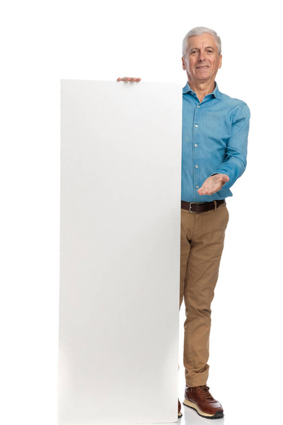 proud man in his 60s with empty board smiling and presenting with his hand while standing and posing on white background - Valokuva, kuva