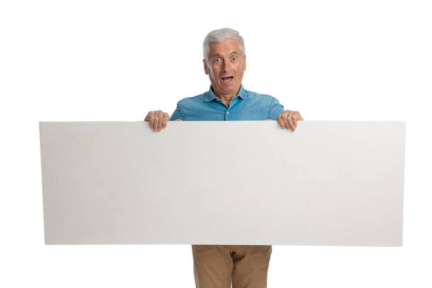 έκπληκτος γέρος κρατώντας άδειο λευκό πίνακα και δείχνει ενώ στέκεται και παρουσιάζει σε λευκό φόντο στο στούντιο - Φωτογραφία, εικόνα