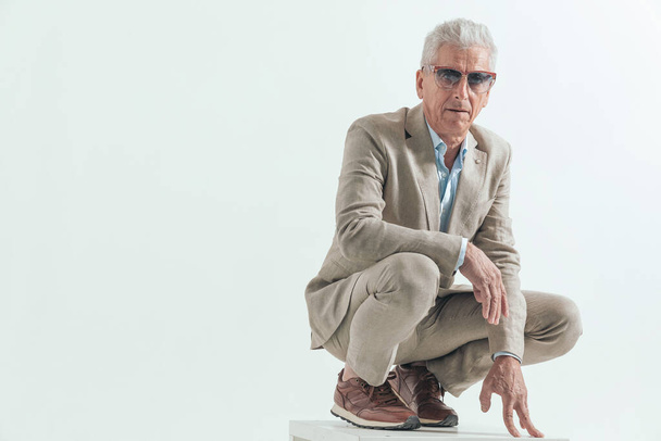 anciano con gafas de sol siendo cool, agachado y sosteniendo los brazos en pose de moda frente a fondo gris  - Foto, imagen