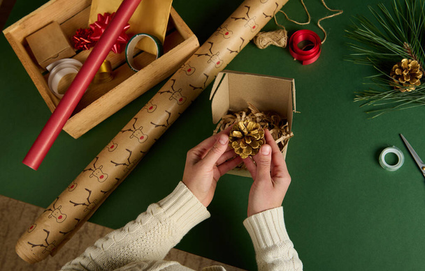 Pohled shora ženy ruce uvedení zlaté borovice kužel do řemeslné lepenkové krabice při balení vánoční dárky. Balení diy dárky. Příprava na Nový rok. Slavnostní zimní událost. Ruční umělecké řemeslo - Fotografie, Obrázek