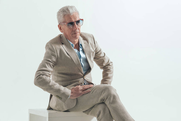 homme de mode cool avec les jambes croisées de cheveux gris, regardant loin et posant d'une manière confiante sur fond gris, assis dans le studio - Photo, image