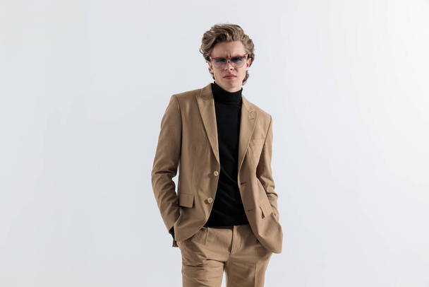 confident young man with blond long hair posing with hands in pockets while standing in front of grey background in a nice beige suit  - Foto, afbeelding