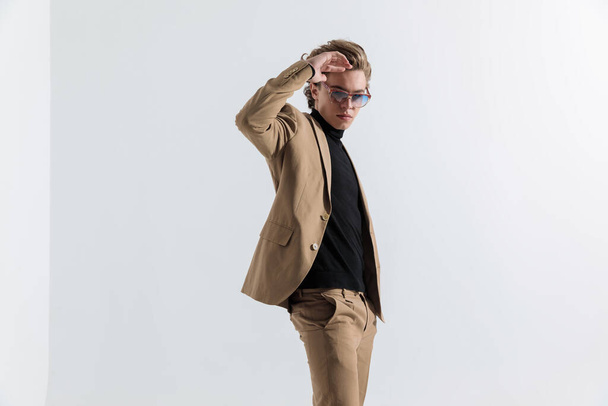 side view of cool guy with sunglasses holding hand to forehead in a fashion pose while standing in front of grey background in studio - Photo, Image