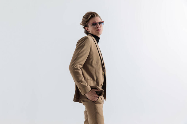 side view of good looking young man with long hair and sunglasses posing with hands in pockets in front of grey background in studio - 写真・画像