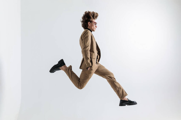 side view of man with long hair jumping in the air with hands in pockets in front of grey background in studio - Photo, image