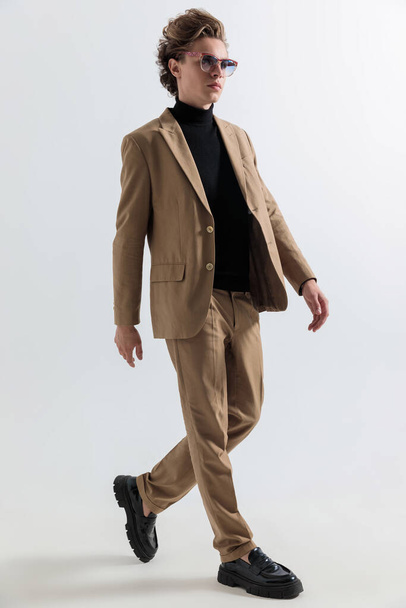jovem homem de moda em roupa casual inteligente olhando para longe e andando na frente de fundo cinza em estúdio - Foto, Imagem