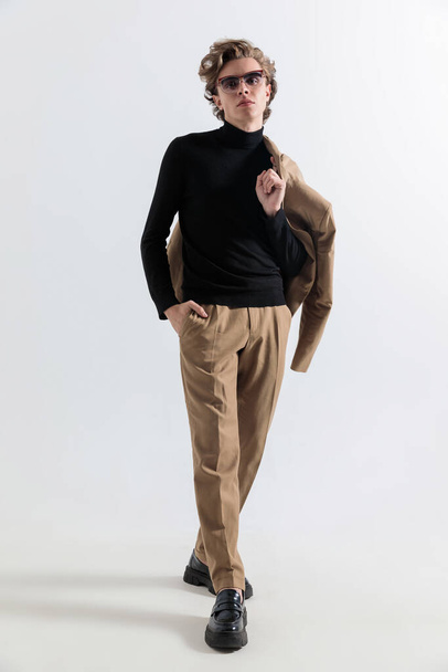 модный мужчина с длинными волосами, который держит пиджак через плечо и ходит с рукой в карманах перед серым фоном в студии - Фото, изображение