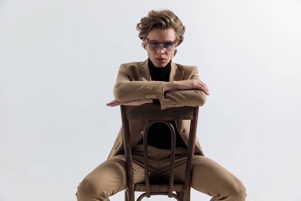 sexy hombre de la moda sosteniendo brazos. en el respaldo de la silla de madera, tocando la frente con la mano y pensando y posando sobre fondo gris en el estudio - Foto, Imagen