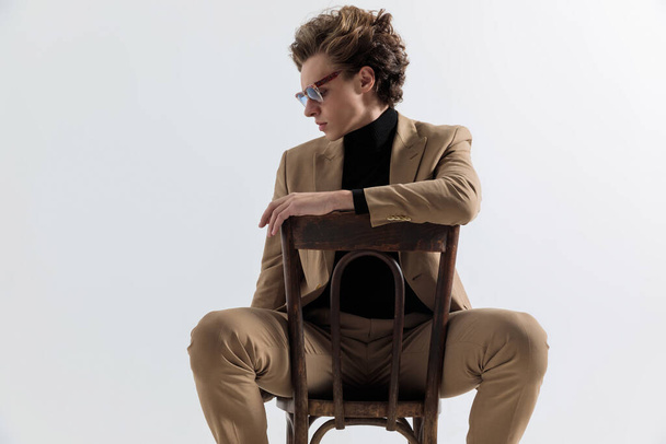 cool jeune homme avec les cheveux longs tenant les bras sur la chaise et regardant vers le bas tout en posant devant fond gris en studio - Photo, image