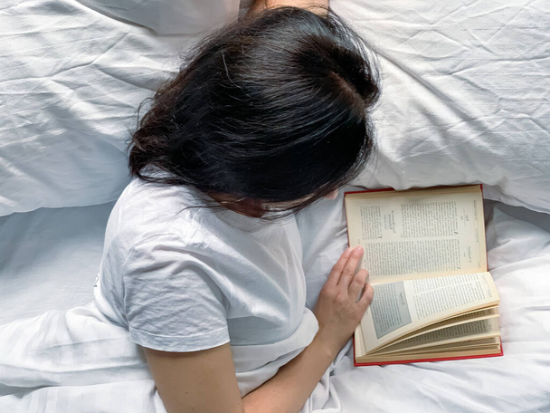 Young millennial candid woman reading red book at home on white striped bedsheets. Early morning. Text in book is blurred. Education or slow living concept. Young adult beautiful woman. High quality - Photo, Image