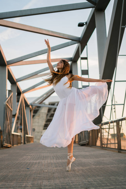 Young adult ballerina dancing on a bridge wearing white dress. - Foto, imagen
